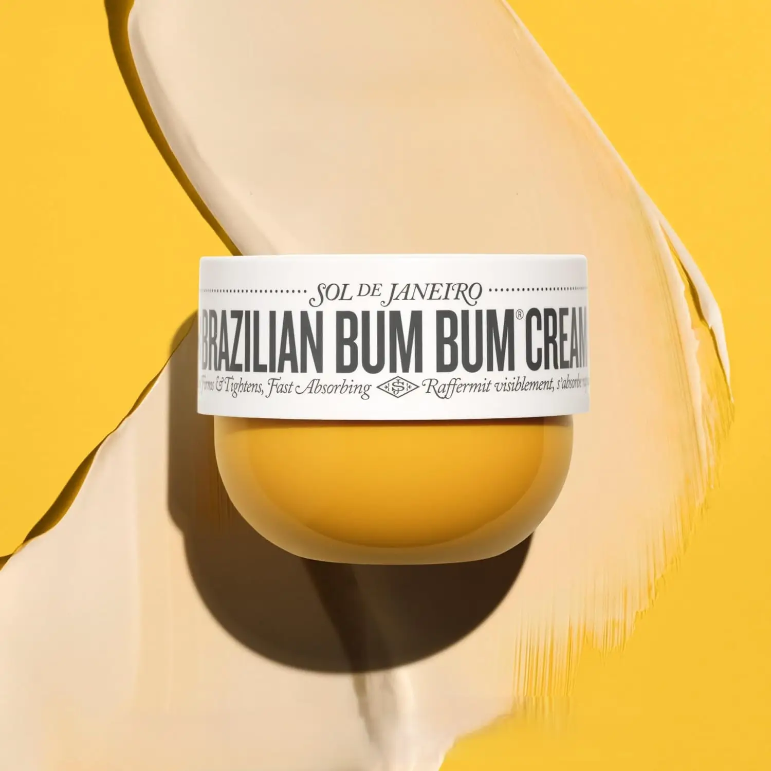 pote original do Sol de Janeiro Brazilian Bum Bum Cream sobre fundo amarelo, com textura cremosa espalhada; hidratante corporal perfumado Cheirosa 62